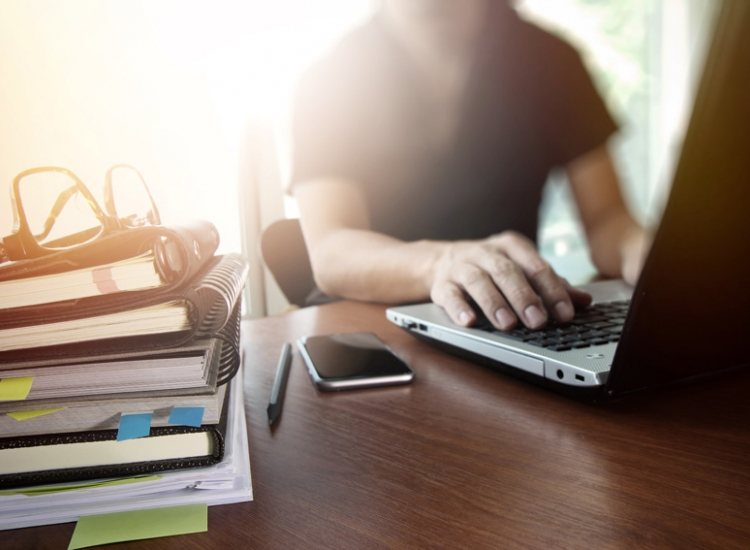Persoon typt op laptop met stapel documenten op bureau