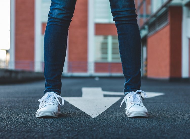 Persoon met witte sneakers op een pijlpunt op de weg, met een rood gebouw op de achtergrond.